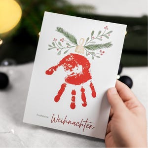 Puede incluir: Tarjeta navideña blanca con una huella de mano roja en forma de adorno navideño, adornada con ramitas verdes y bayas rojas. La tarjeta tiene la inscripción "Fröhliche Weihnachten" en escritura roja.