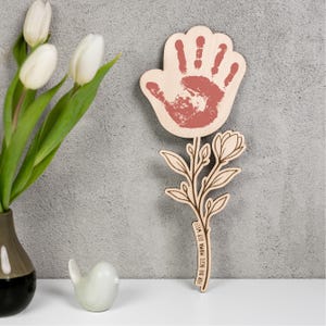 Könnte beinhalten: Holzblumenförmige Dekoration mit einem Handabdruck in der Mitte. Der Handabdruck ist rotbraun. Der Stiel hat ein Blumendesign und den Text "FOR THE BEST MOM IN THE WORLD".