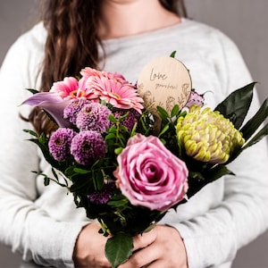 Könnte beinhalten: Ein lebendiger Blumenstrauß mit rosa Gerbera, lila Calla-Lilien und einer großen rosa Rose. Ein Holzschild mit der Aufschrift "Love grows here!" ist zwischen den Blüten platziert. Der Strauß wird von einer Person in einem grauen Pullover gehalten.