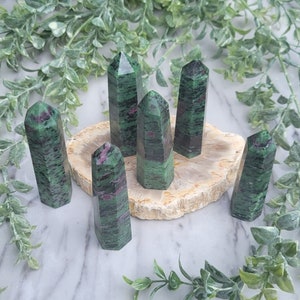 May include: Six green and pink gemstone points, also known as green ruby, are arranged on a light brown stone base. The points are all approximately the same size and shape, and they have a smooth, polished finish.