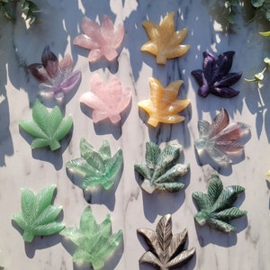 May include: A collection of 14 carved crystal leaf shapes in various colors, including green, pink, purple, and yellow. The leaves are arranged on a white marble surface.