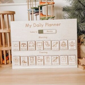 May include: A wooden daily planner with a light wood finish. The planner has a section for "Morning" and "Evening" activities. Each section has a row of eight icons representing different activities, such as "wake up", "feed dogs", "breakfast", "screen time", "get dressed", "brush teeth", "pack bag", "school", "dinner", "tidy up", "use toilet", "bath time", "pyjamas on", "brush teeth", "story time", and "bed time". The planner also has a section for "Today is:" and a blank space for the day of the week. The day of the week is "Monday".