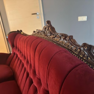 Puede incluir: Un sofá de terciopelo rojo con tallas de madera ornamentadas y detalles dorados. El sofá tiene un respaldo capitoné y un diseño clásico.