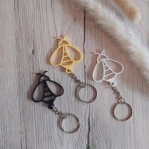 May include: Three bee-shaped keychains in yellow, white, and black. Each keychain features a detailed bee design with a chain and a silver-colored key ring. The keychains are displayed on a light wood-grain surface.
