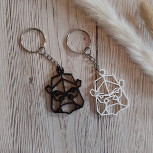 May include: Two keychains featuring geometric gorilla designs. One keychain is black, the other is white. Both have silver key rings and chain attachments. The keychains are displayed on a light wood surface with decorative elements.