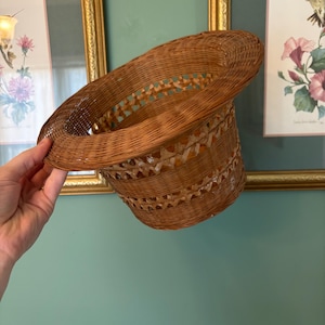 May include: A brown woven wicker hat with a wide brim. The hat is held up against a light teal wall, with framed pictures in the background. The hat has a decorative woven pattern around the crown.