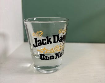 May include: A clear shot glass with the words "Jack Daniel's Old No. 7" in black and gold lettering. The shot glass is on a white surface with a green background.