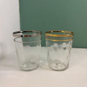 May include: Two clear glass tumblers with decorative rims. One tumbler has a silver rim and a silver band around the middle. The other tumbler has a gold rim and a textured pattern on the glass. Both glasses are empty.