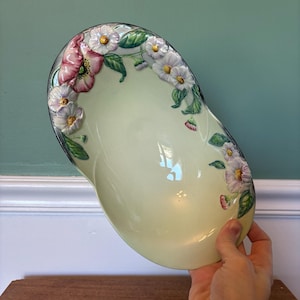 May include: A pale green oval serving dish with a raised floral design. The flowers are white with pink centers and green leaves. The dish is made of ceramic.