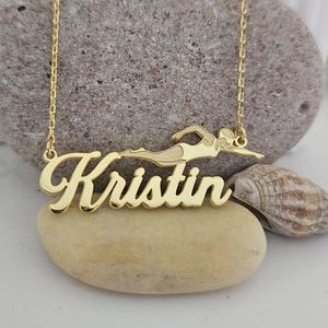 Peut inclure: Collier doré avec le prénom "Kristin" en écriture cursive, accompagné d'une silhouette de nageur. Le pendentif est suspendu à une chaîne délicate. Le collier est présenté sur une pierre claire.