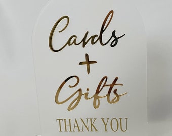 May include: A white acrylic sign with gold lettering that reads "Cards + Gifts Thank You".