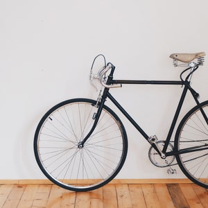 May include: A black bicycle with a brown leather saddle, thin tires, and a white wall background.