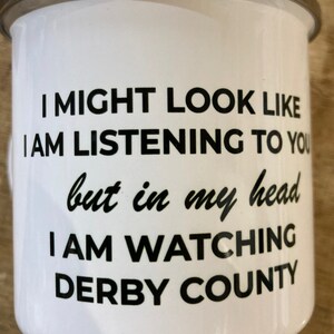 Op de afbeelding: Een witte emaille mok met zwarte tekst die luidt: "Ik zie er misschien uit alsof ik luister, maar in mijn hoofd kijk ik Derby County".