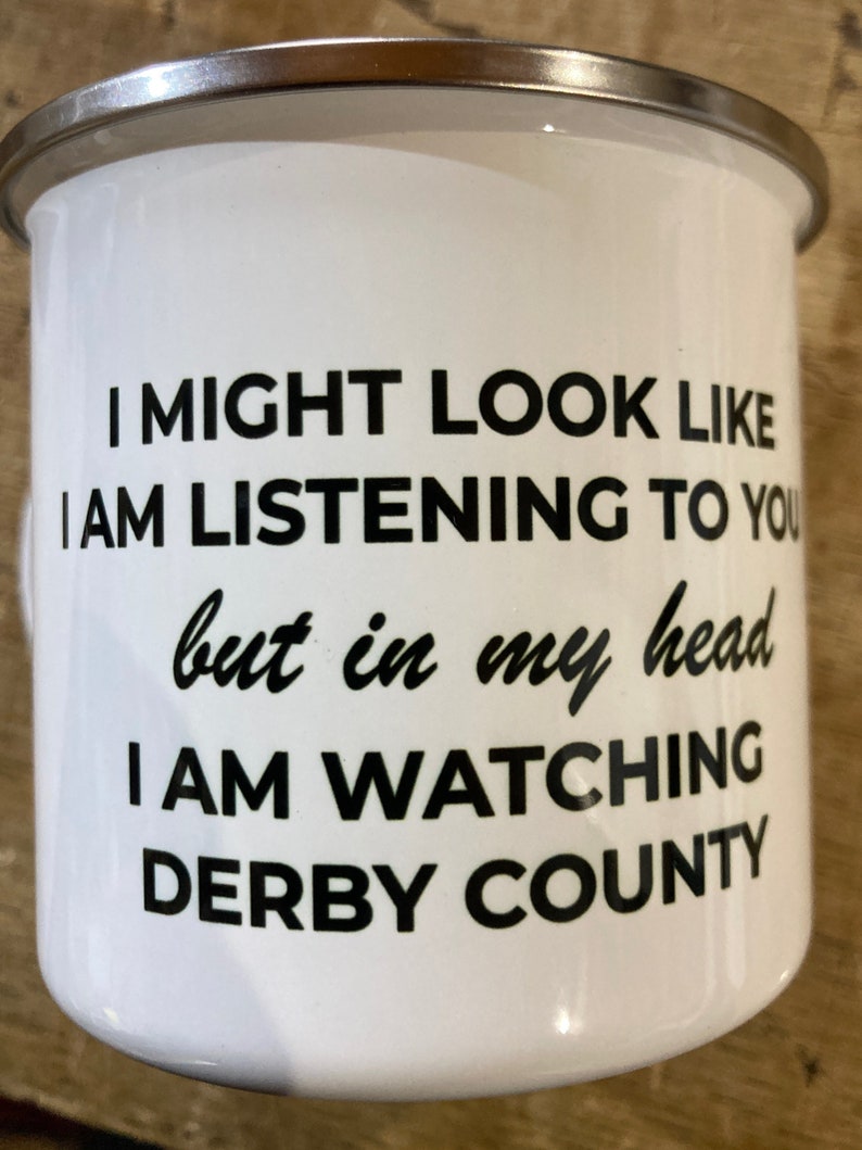 Op de afbeelding: Een witte emaille mok met zwarte tekst die luidt: "Ik zie er misschien uit alsof ik luister, maar in mijn hoofd kijk ik Derby County".