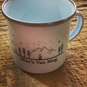 Pu&ograve; includere: Tazza da campeggio in smalto bianco con un design a silhouette nera di montagne, alberi e un fal&ograve;. Il testo "Mike's Van Mug" &egrave; stampato sulla tazza.