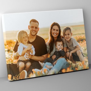 May include: A family of five pose for a photograph in a field of yellow flowers. The parents are sitting on the ground with their three children. The father is wearing a black shirt and the mother is wearing a blue shirt. The children are wearing a variety of colours.