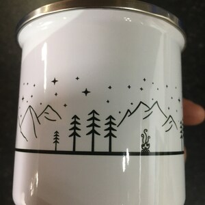 Pu&ograve; includere: Tazza da campeggio in smalto bianco con un design a silhouette nera di montagne, alberi e un fal&ograve; sotto un cielo stellato.