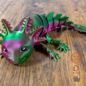 Puede incluir: Una figura de axolotl impresa en 3D con un acabado iridiscente verde y rosa. La figura tiene un cuerpo articulado detallado y una expresión amigable.