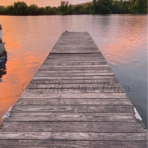 May include: A wooden dock extends into calm water, reflecting the colors of a sunset sky. The dock is made of weathered planks, with a cleat visible on the side. Trees line the distant shoreline, creating a serene waterscape.