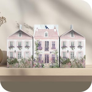 Pode incluir: Três casas rosa em miniatura com molduras e janelas brancas, cercadas por folhagem verde e flores roxas. Um pequeno pássaro azul pousa no telhado da casa do meio.