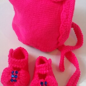 May include: A pink knitted baby hat with a bow and matching booties. The hat has a long tie and both the hat and booties have blue rhinestone embellishments.