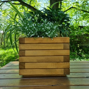 May include: A wooden planter box with a light brown stain, filled with green plants and white flowers. The planter is sitting on a wooden table.