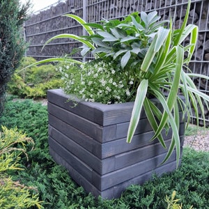 May include: A dark gray wooden planter box with a variety of green plants growing inside. The planter is made of wood planks and has a rustic look.