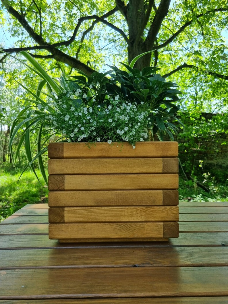 May include: A wooden planter box with a light brown stain, filled with green plants and white flowers. The planter is sitting on a wooden table.
