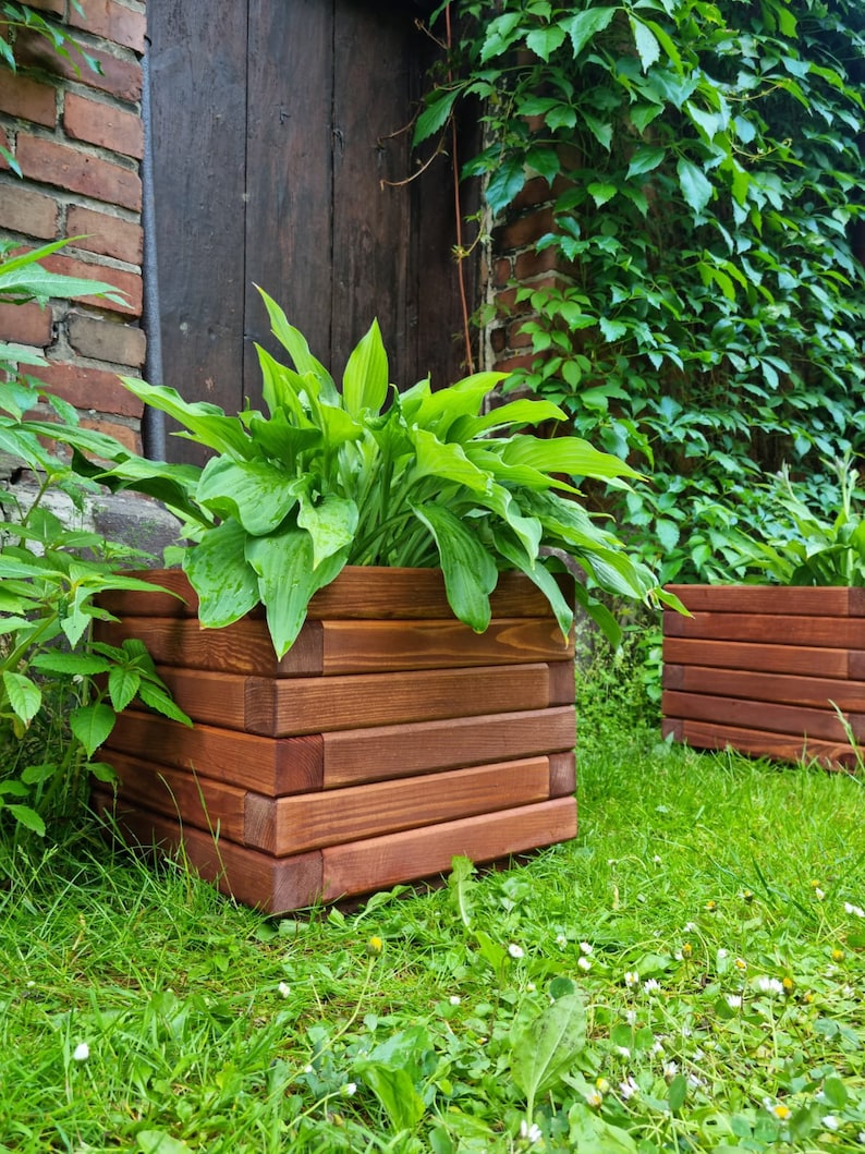May include: Two wooden planter boxes with a dark brown stain. The planter boxes are filled with green plants and are sitting on a bed of green grass.