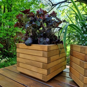 May include: Two wooden planter boxes with plants. The planter boxes are made of stacked wooden planks and are a light brown color. The planter boxes are sitting on a wooden deck.