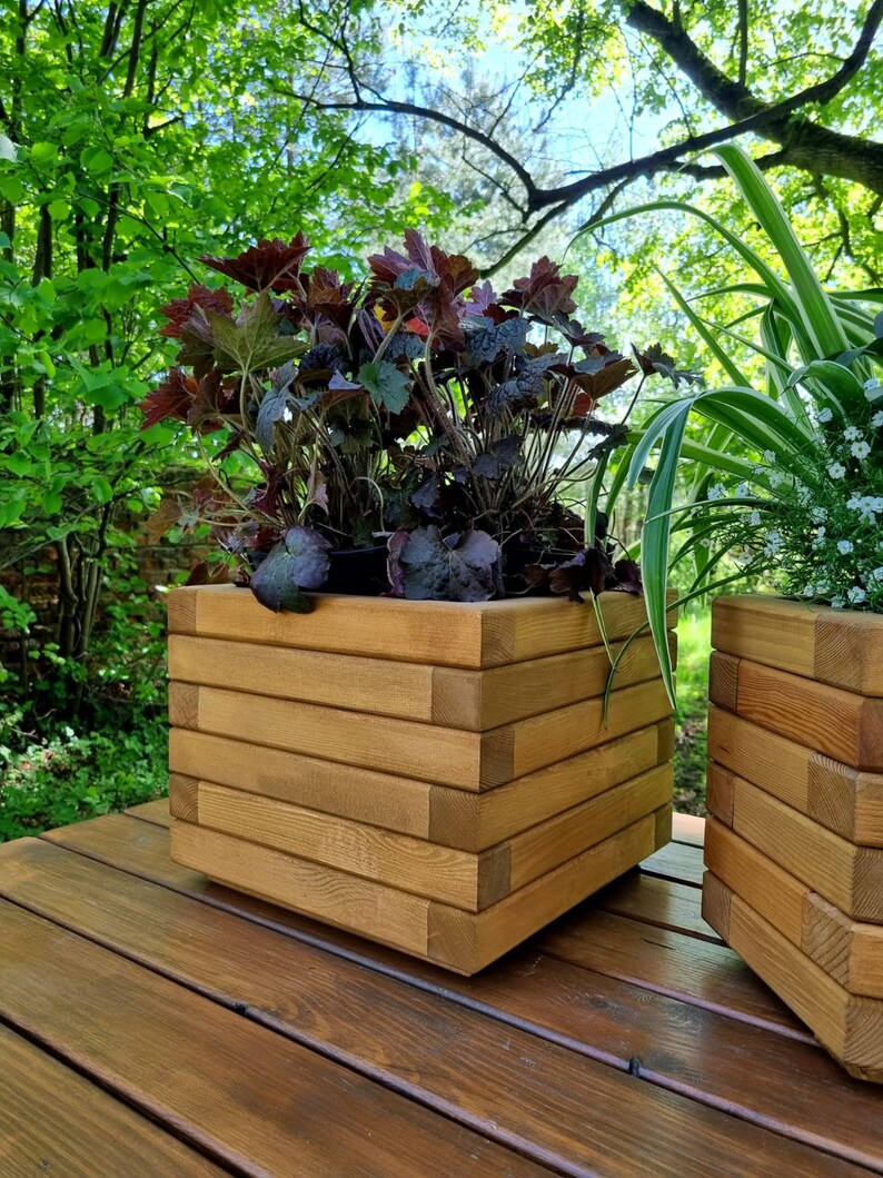 May include: Two wooden planter boxes with plants. The planter boxes are made of stacked wooden planks and are a light brown color. The planter boxes are sitting on a wooden deck.