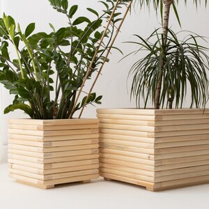Op de afbeelding: Twee houten plantenpotten met een natuurlijke afwerking. De potten zijn gemaakt van gestapelde houten latten en zijn vierkant. De potten zijn leeg en staan op een wit oppervlak.