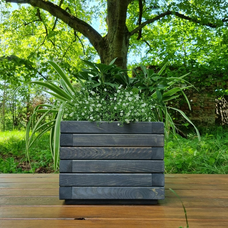 May include: A dark gray wooden planter box with a variety of green plants and white flowers. The planter is sitting on a wooden surface.