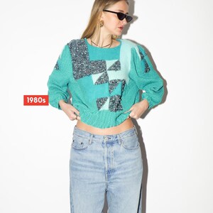 Può includere: Un maglione turchese con motivi geometrici nelle tonalità del turchese e del grigio. Il maglione ha maniche lunghe e scollo tondo. Il testo "1980s" è in un riquadro rosso in basso a sinistra. Il maglione è abbinato a jeans in denim chiaro.