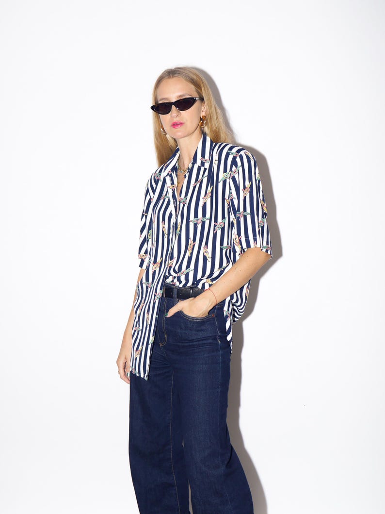 May include: A short-sleeved, button-down shirt with vertical navy blue and white stripes. The shirt features a pattern of small, colorful designs. Paired with dark wash, wide-leg jeans and a black belt.