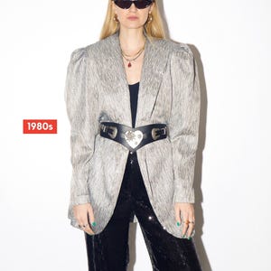 Op de afbeelding: Een zilverkleurige blazer met schoudervullingen, gecombineerd met een zwarte riem met een hartvormige gesp en een zwarte paillettenbroek. De afbeelding bevat de tekst "1980s" in een rode doos, wat wijst op een vintage modethema.