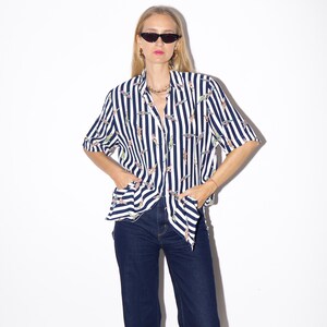 May include: A short-sleeved button-down shirt with vertical navy blue and white stripes. The shirt features a repeating pattern of colorful, abstract shapes. The shirt is paired with dark blue denim jeans.
