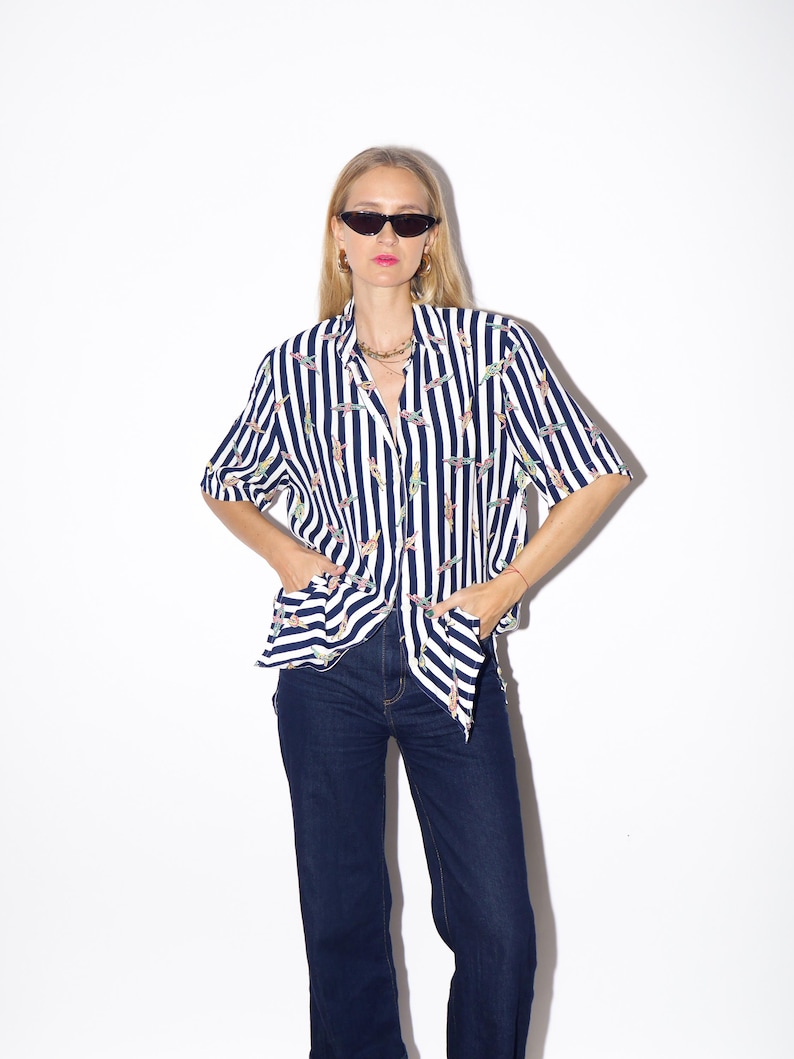 May include: A short-sleeved button-down shirt with vertical navy blue and white stripes. The shirt features a repeating pattern of colorful, abstract shapes. The shirt is paired with dark blue denim jeans.