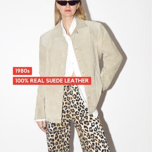 Pu&ograve; includere: Giacca in camoscio beige chiaro con chiusura a bottoni, abbinata a pantaloni con stampa leopardata. L'immagine include il testo "1980s" e "100% REAL SUEDE LEATHER". La giacca &egrave; in stile vintage.