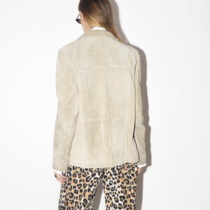 Pu&ograve; includere: Giacca in pelle scamosciata color beige chiaro con collo e chiusura con bottoni. La giacca &egrave; abbinata a pantaloni con stampa leopardata. La modella indossa occhiali da sole e una camicia bianca sotto la giacca.