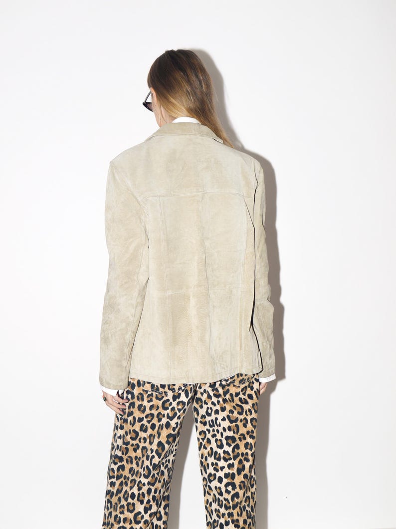 Pu&ograve; includere: Giacca in pelle scamosciata color beige chiaro con collo e chiusura con bottoni. La giacca &egrave; abbinata a pantaloni con stampa leopardata. La modella indossa occhiali da sole e una camicia bianca sotto la giacca.