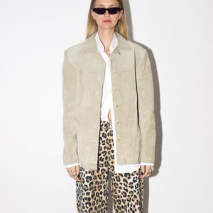 Pu&ograve; includere: Giacca in camoscio beige chiaro con bottoni, indossata sopra una camicia bianca con colletto e pantaloni con stampa leopardata. La giacca ha un design classico e su misura. I pantaloni hanno un motivo maculato marrone e nero.