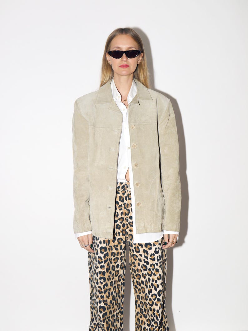Pu&ograve; includere: Giacca in camoscio beige chiaro con bottoni, indossata sopra una camicia bianca con colletto e pantaloni con stampa leopardata. La giacca ha un design classico e su misura. I pantaloni hanno un motivo maculato marrone e nero.
