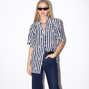 May include: A short-sleeved, button-down shirt with vertical navy blue and white stripes. The shirt features a repeating pattern of colorful figures. The shirt is paired with dark blue jeans and a black belt.