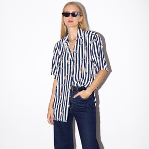 May include: A short-sleeved button-down shirt with vertical navy blue and white stripes. The shirt features a pattern of colorful, abstract shapes. The shirt is paired with dark wash, wide-leg jeans. The model is wearing black sunglasses.