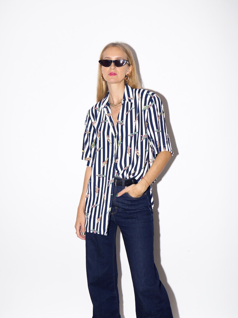 May include: A short-sleeved button-down shirt with vertical navy blue and white stripes. The shirt features a pattern of colorful, abstract shapes. The shirt is paired with dark wash, wide-leg jeans. The model is wearing black sunglasses.