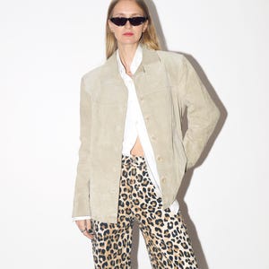 Pu&ograve; includere: Giacca in camoscio beige chiaro con bottoni, indossata sopra una camicia bianca con colletto. L'outfit comprende pantaloni con stampa leopardata e occhiali da sole neri. La giacca ha un design classico e su misura.