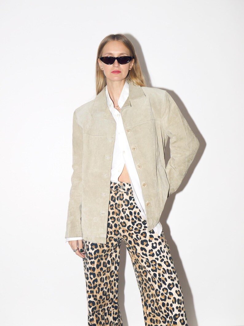 Pu&ograve; includere: Giacca in camoscio beige chiaro con bottoni, indossata sopra una camicia bianca con colletto. L'outfit comprende pantaloni con stampa leopardata e occhiali da sole neri. La giacca ha un design classico e su misura.