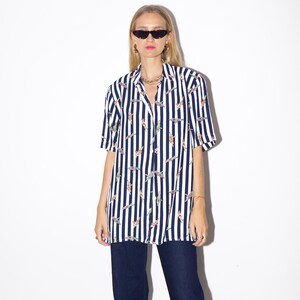 May include: A short-sleeved, button-down shirt with vertical navy blue and white stripes. The shirt features a pattern of colorful figures. The shirt is paired with dark blue jeans. The model is wearing sunglasses and jewelry.