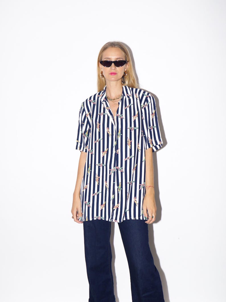 May include: A short-sleeved, button-down shirt with vertical navy blue and white stripes. The shirt features a pattern of colorful figures. The shirt is paired with dark blue jeans. The model is wearing sunglasses and jewelry.